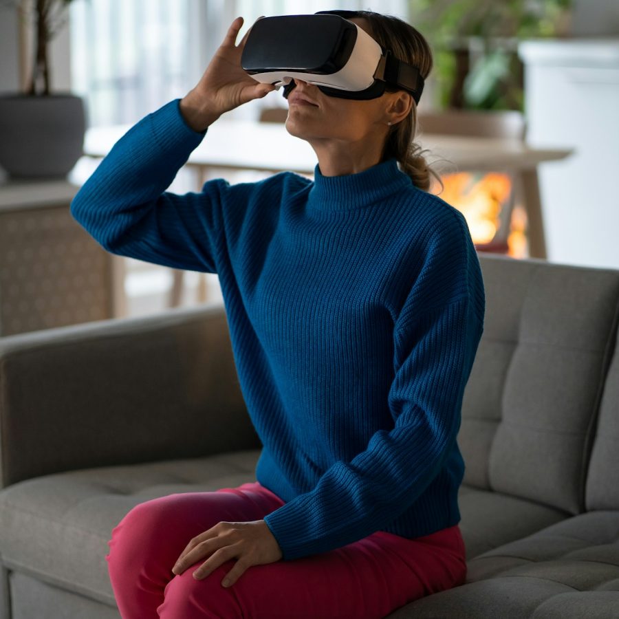 Carried away European woman in virtual reality headset looking up using VR and AR sits on sofa